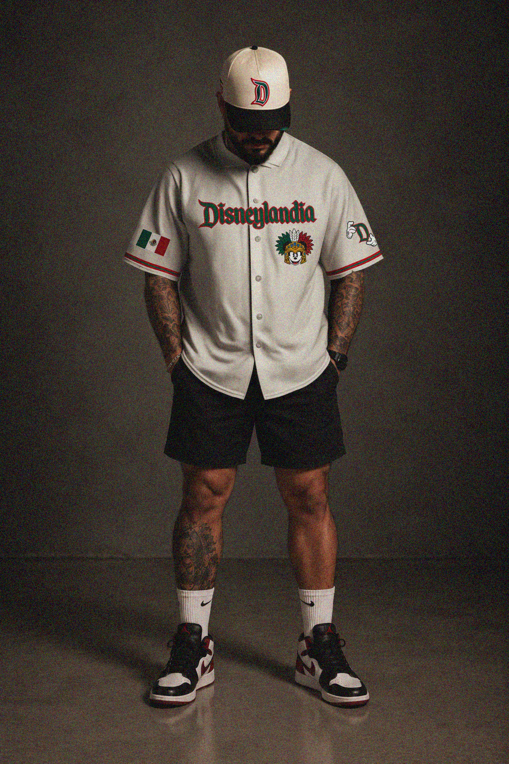 A man stands against a dark studio background wearing a white Disneyland baseball-style jersey with black shorts, white socks, and black-and-white sneakers. The jersey has 'Disneylandia' across the chest, a multicolored character patch, and a Mexican flag patch on the sleeve.
